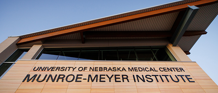 Front view of Munroe-Meyer Institute, looking up at black lettered signage on tan exterior building and rust-colored angled roof, blue sky behind; Credit UNMC photographer, Kent Sievers 2020.