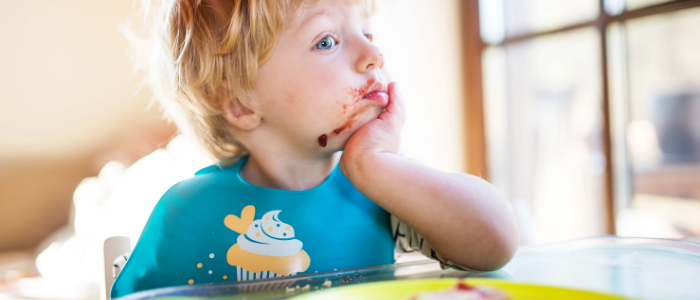stock photo of young boy wearing a bib with a birthday cupcake printing, sitting at a table, one elbow on table top, hand propped under chin, face messy with food; credit: Canva
