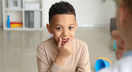 stock photo of young boy with hand touching mouth and lips, looking at therapist, during a speech-therapy session; credit: Adobe
