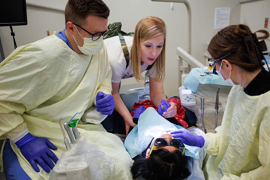 Two dental students clean a child's teeth while a social worker holds her hands.