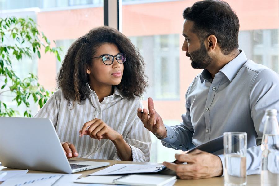 A man and woman sit at a desk in conversation with a laptop and papers before them