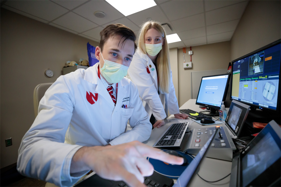 Two people in white lab coats look at a computer monitor.