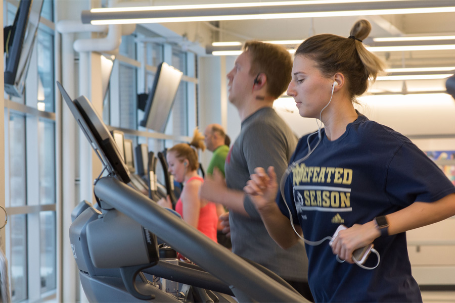 A woman wearing headphones runs on a treadmill with other runners in the background