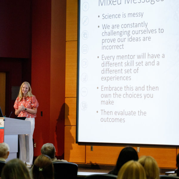 a woman at a podium talking to crowd with slides behind her