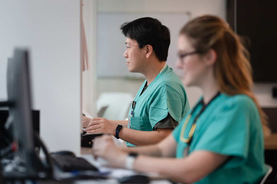 Two students in teal scrubs work at computers