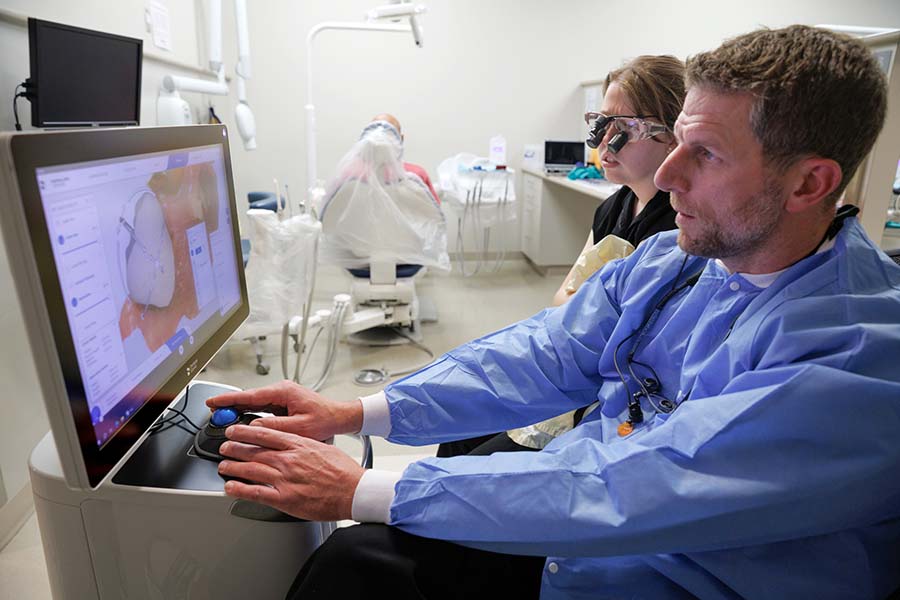 A student and faculty member work on a case plan on a screen while a patient waits in the background.