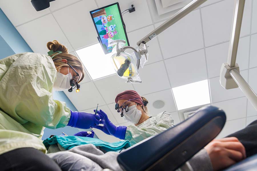 Two female residents wear dental loupes while passing an instrument and caring for a patient with a cartoon playing on a ceiling mounted monitor.