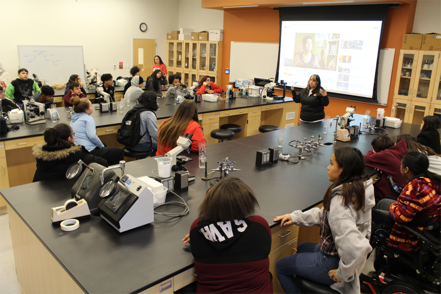 A lecturer speaks to a classroom of middle school students sitting at tables in a learning laboratory.