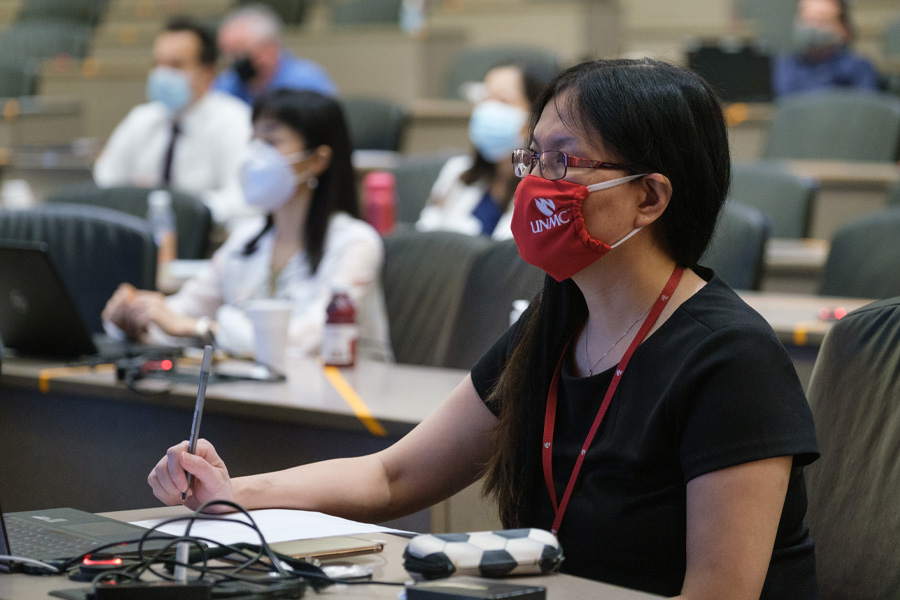 A woman wearing glasses and a surgical mask listens to a lecturer out of frame.