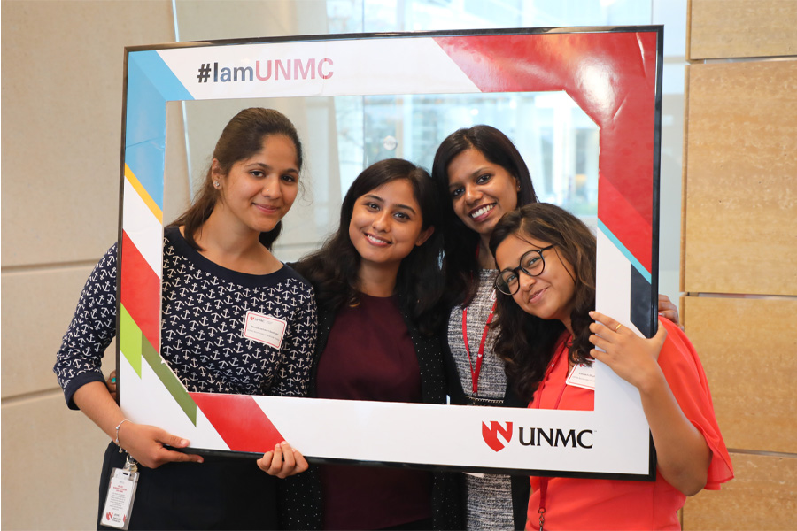 Four women pose for a picture in a cardboard picture frame with the words "#IamUNMC" and the UNMC logo.