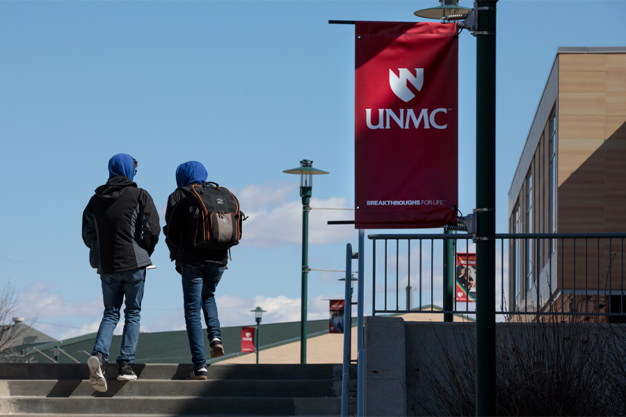 Two students wearing backpacks walk past a banned with the UNMC logo.