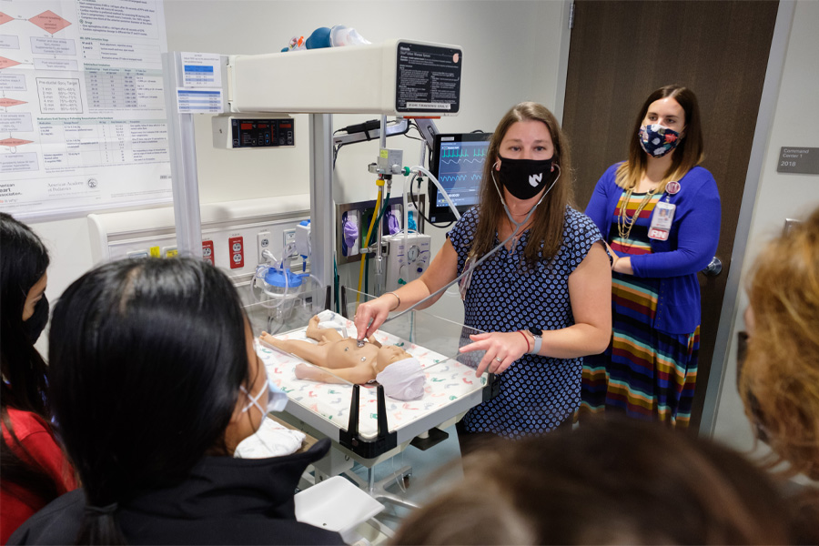 A woman wearing a cloth mask teaches a lesson to a group of students using a model of an infant