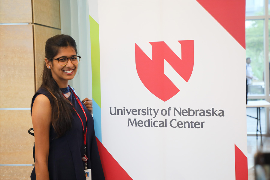 A woman wearing glasses poses next to a banner with the UNMC logo