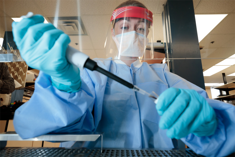 A woman wearing a surgical mask and face shield uses a pipette in a lab.