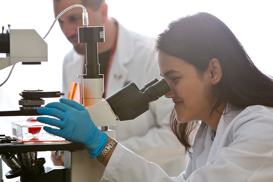 Student looks as slide in a microscope while faculty member talks to her.