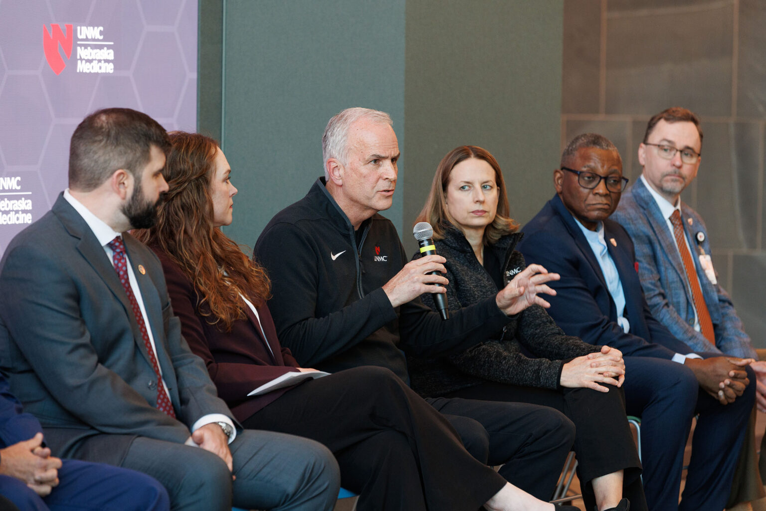 Panel discussion with multiple professionals seated on stage, one speaking into a microphone at a UNMC and Nebraska Medicine event.