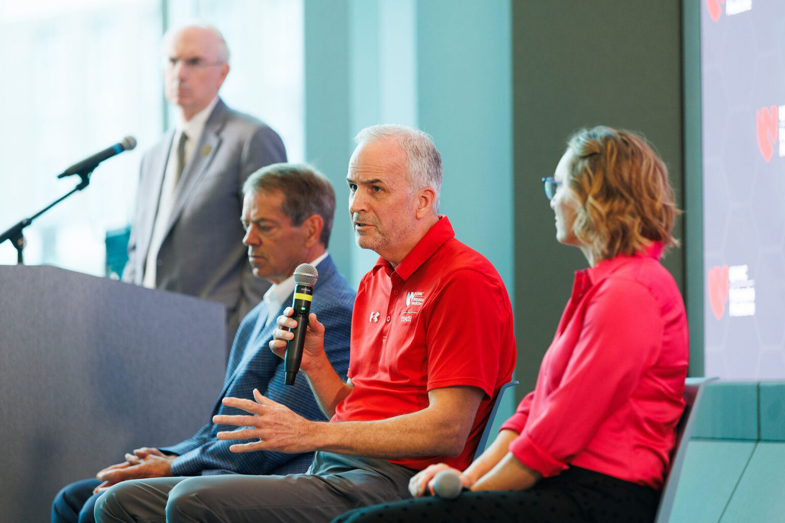 Panel discussion with a speaker in a red shirt holding a microphone, seated beside two others, with a podium and UNMC branding in the background.