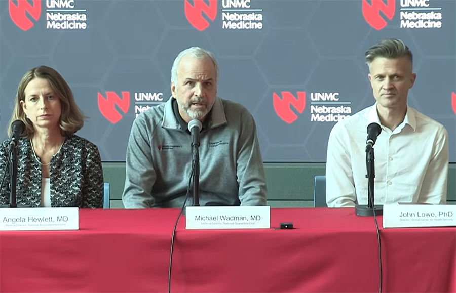 Three individuals seated at a table with microphones during a UNMC/Nebraska Medicine press conference, with a branded backdrop behind them.