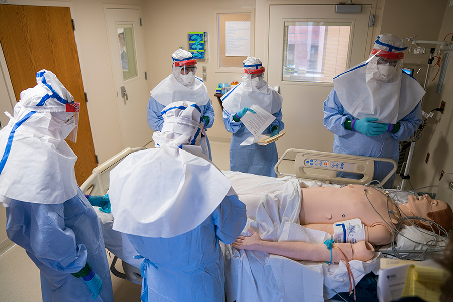 Several people wearing full protective medical gear stand around a hospital bed during a simulated patient care scenario, with a medical training mannequin lying on the bed in a clinical room.