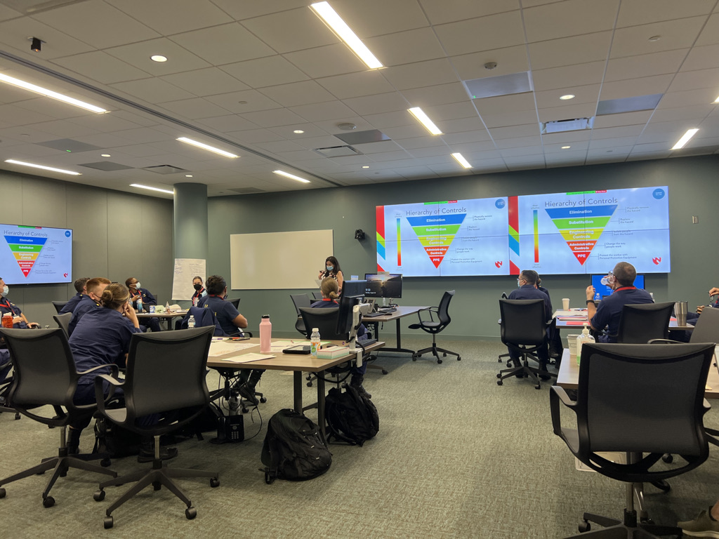 A classroom with attendees in dark uniforms seated at tables, facing a presenter near a screen displaying a "Hierarchy of Controls" chart. The room is well-lit and organized.