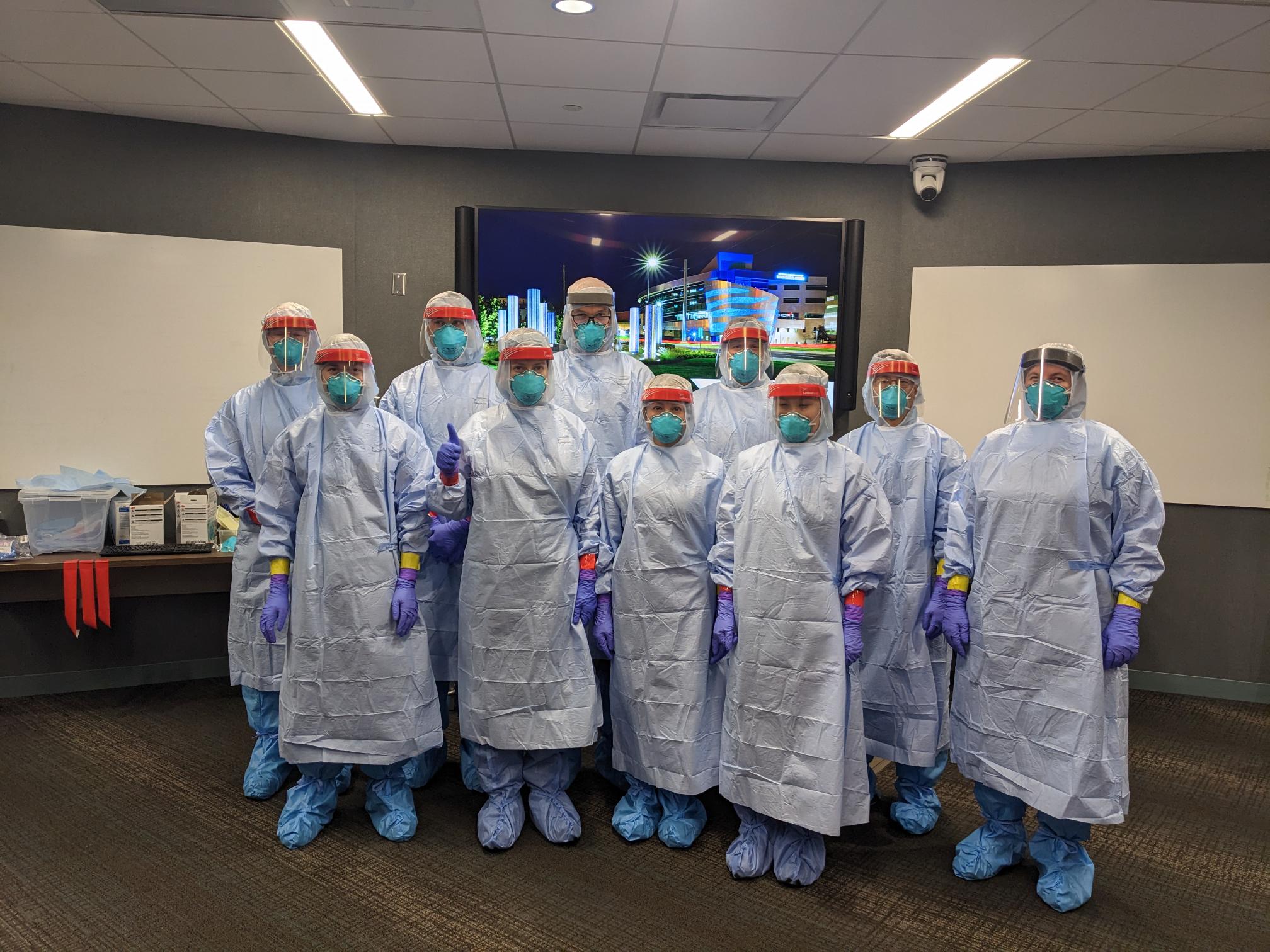 A group of healthcare workers in full protective gear, including face shields, masks, and gloves, stand together in a well-lit room, conveying teamwork and readiness.