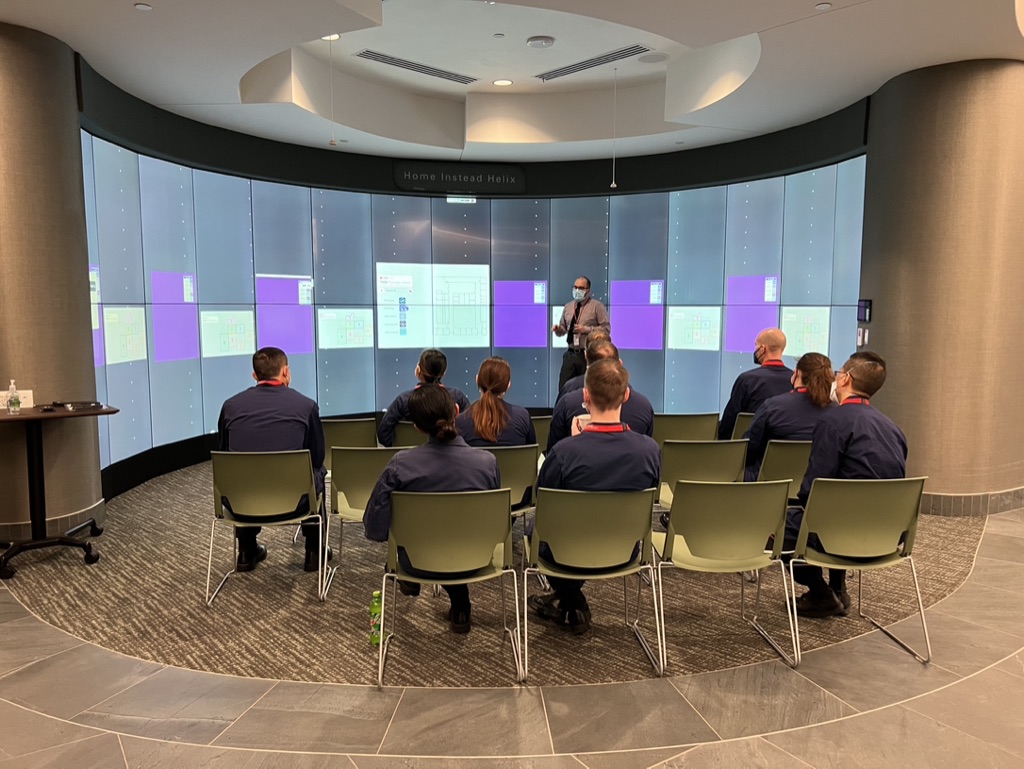 A group of people in matching uniforms sits facing a large screen displaying graphs and charts. A speaker in a mask stands in front, presenting. The setting is modern and professional.