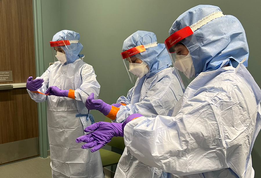 Three people wearing full protective medical clothing, face shields, and gloves stand indoors while practicing donning or adjusting protective gloves.