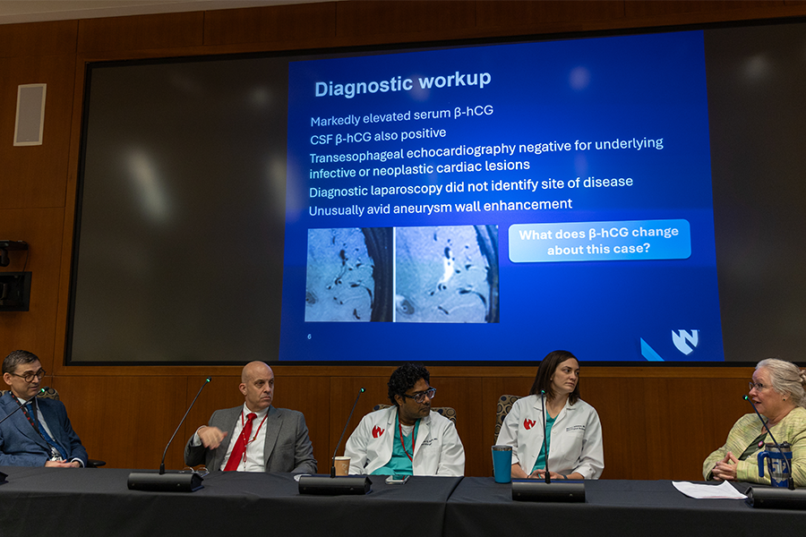 A panel of doctors sit at a long table discussing a case study. Behind them is a presentation of the case study