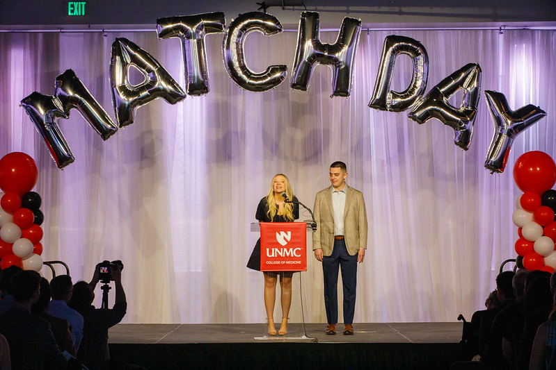 A graduating UNMC medical student announces her residency match on a stage in front of a podium as her significant other looks on.