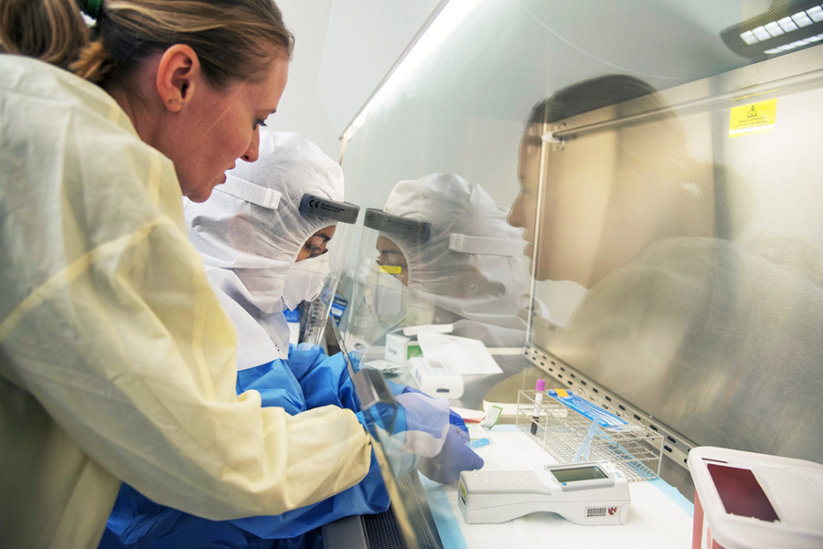 Two UNMC researchers wearing personal protective equipment as they conduct research testing and training in front of a vent hood with test tubes and scientific equipment.