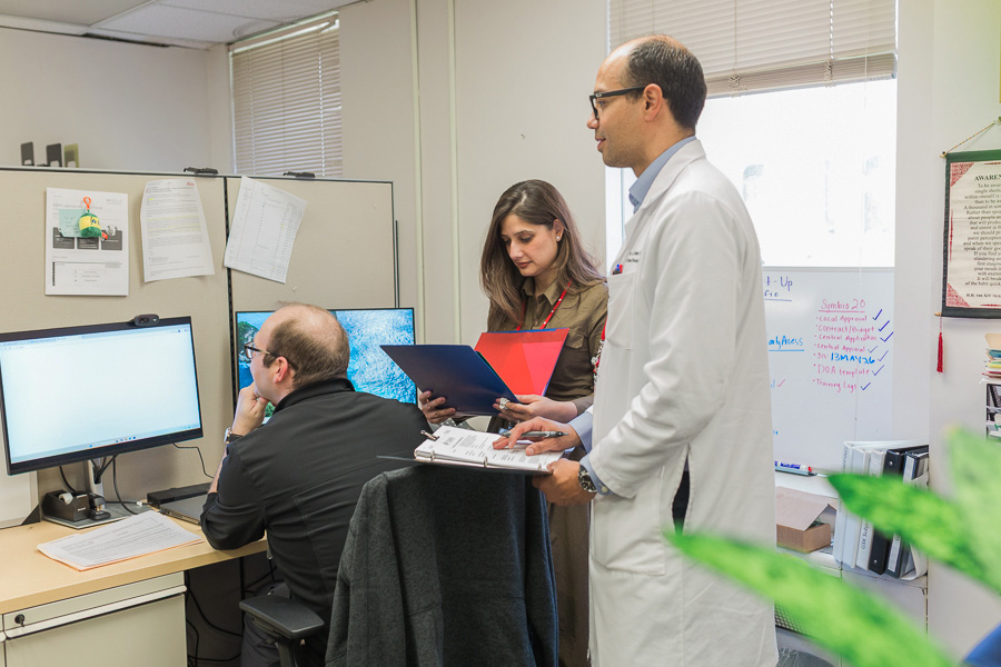 A group of people work together in an office space, two people look at an image on a computer screen while one person reads data in a binder.