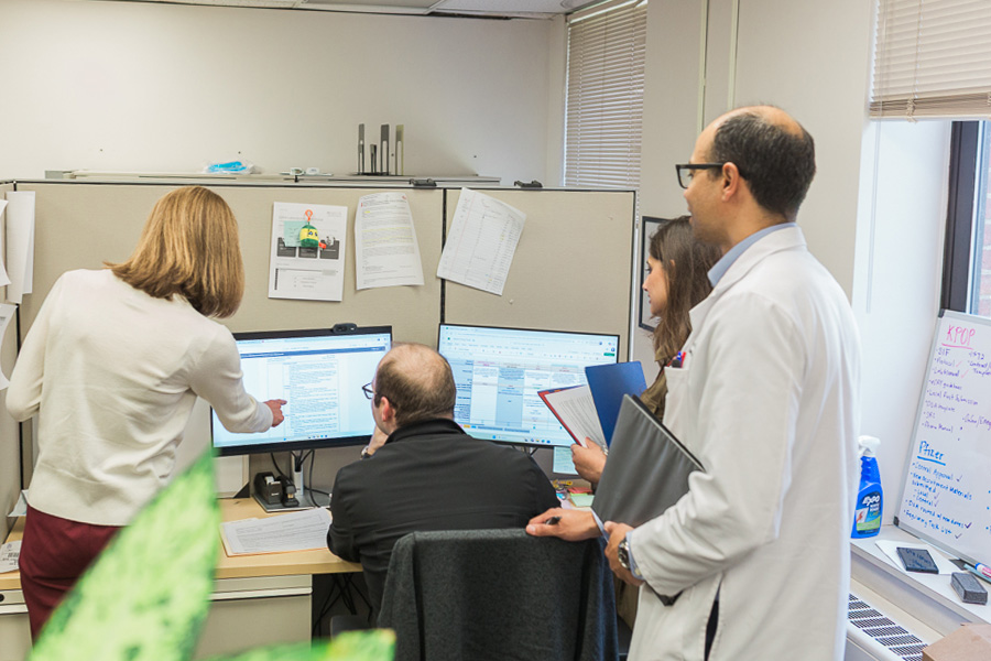 A group of four people work together in an office space and look at an image on screen while one person points at something on screen.