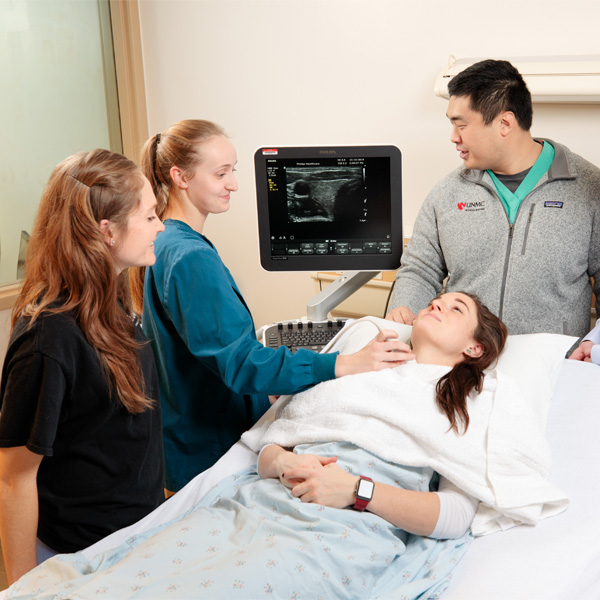 Three students read results from a scan of a patient lying on a bed