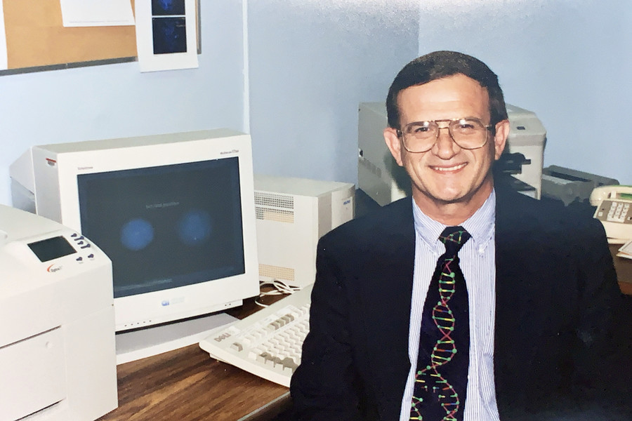 Dr. Sanger sits in front of a computer, facing the camera with a smile, wearing a dark suit and glass, and a DNA-print tie.