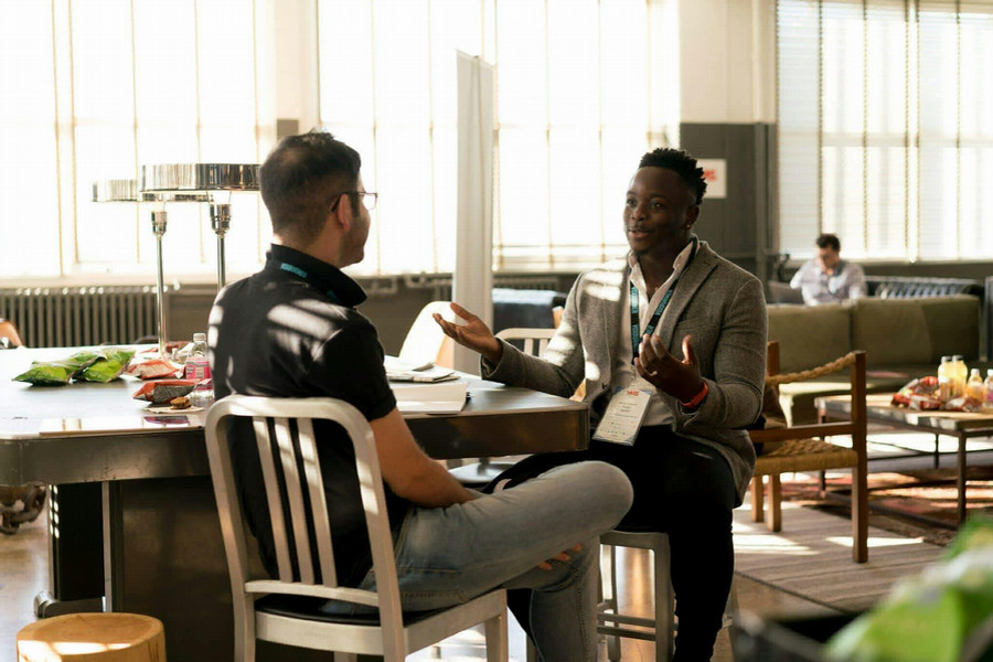 Two individuals converse at a conference table in a sunny room.