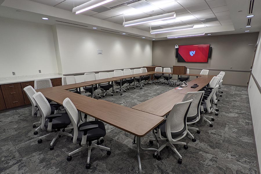 Chairs and tables form a large, open rectangle shape in MMI’s medium conference room, a flat screen TV on one wall.