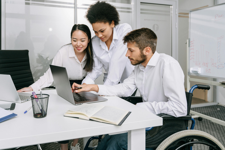 Three individuals, one in a wheelchair, sit at a conference room, working on a laptop.