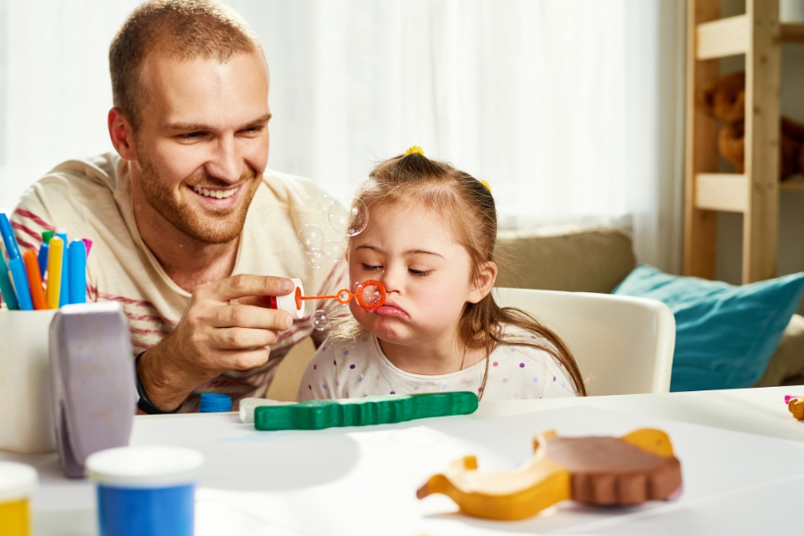 Adult holds up a bubble wand, allowing a small child to blow bubbles.