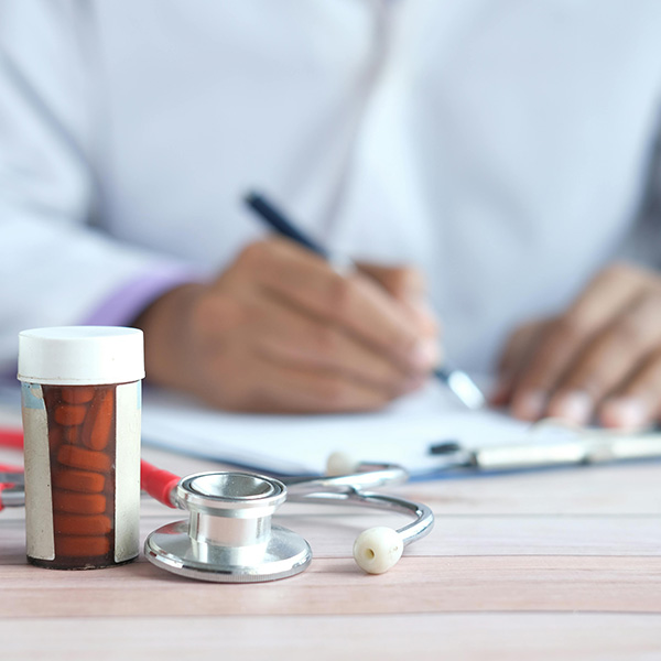 Medication bottles in foreground and medical provider filling out paperwork in the background, credit – Towfiqu Barbhuiya, Pexels.