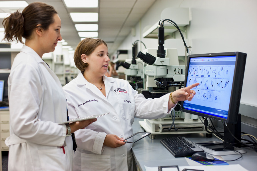 Two women in white lab coats discuss lab findings on a computer.