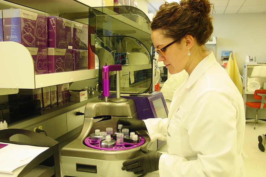 Woman with glasses and white lab coat operates lab testing equipment.