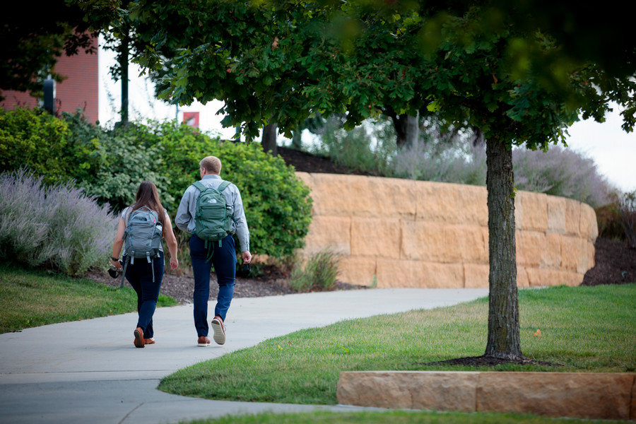 Two students with backpacks walking up a concrete path under a shady tree.