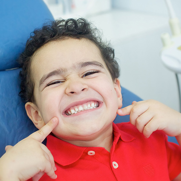 Child in dentist chair pointing at cheeks, smiling, Adobe Stock. 