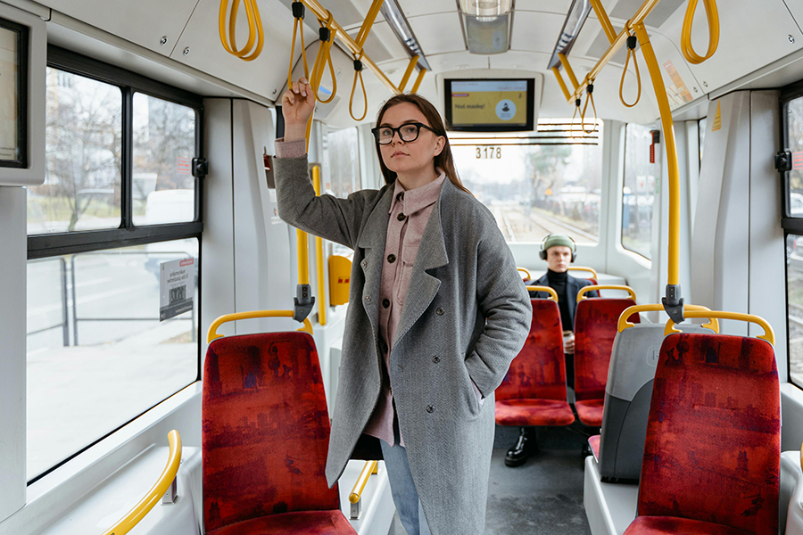 A young adult rides on a city bus, standing in the aisle and holding onto an overhead handle.