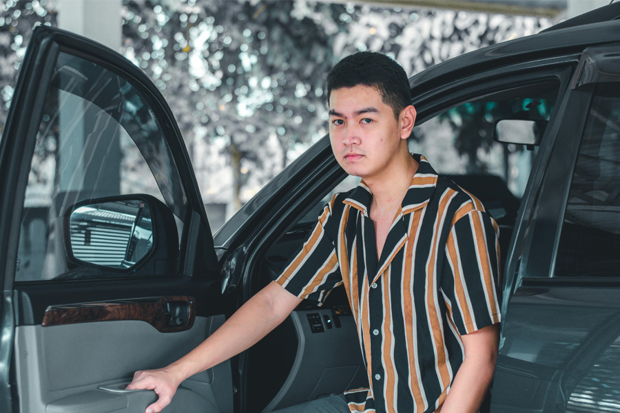 Young man in a striped shirt stands next to a car with the driver side door open.