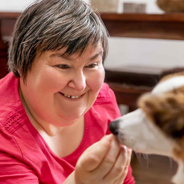 Adult with short dark and gray hair and intellectual disabilities smiles as they feed a dog, Adobe Stock.
