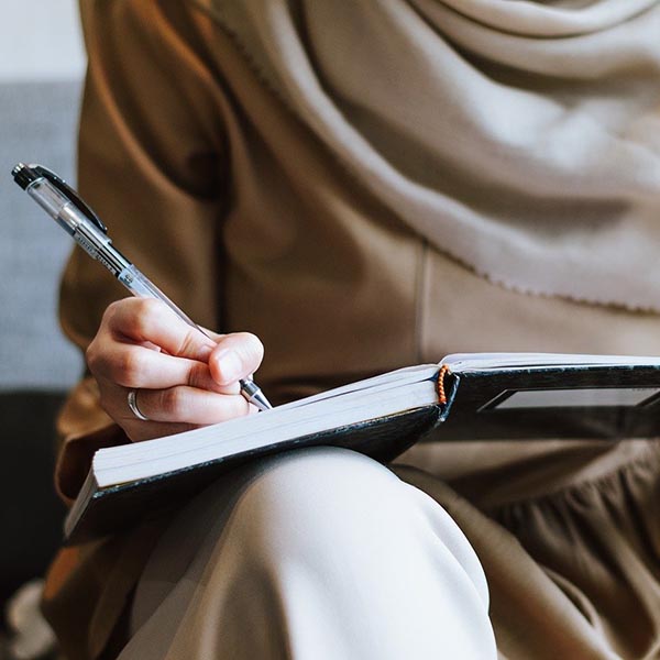 Muslim woman writing in notebook on her lap, by IqbalStock – Pixabay.