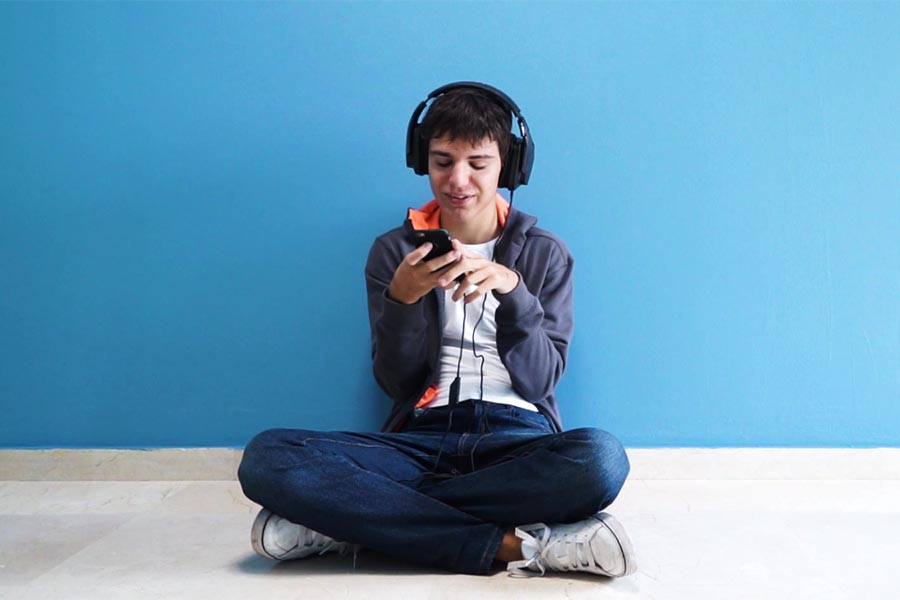 Teen with Autism wearing headphones sitting cross-legged against a wall, Canva.