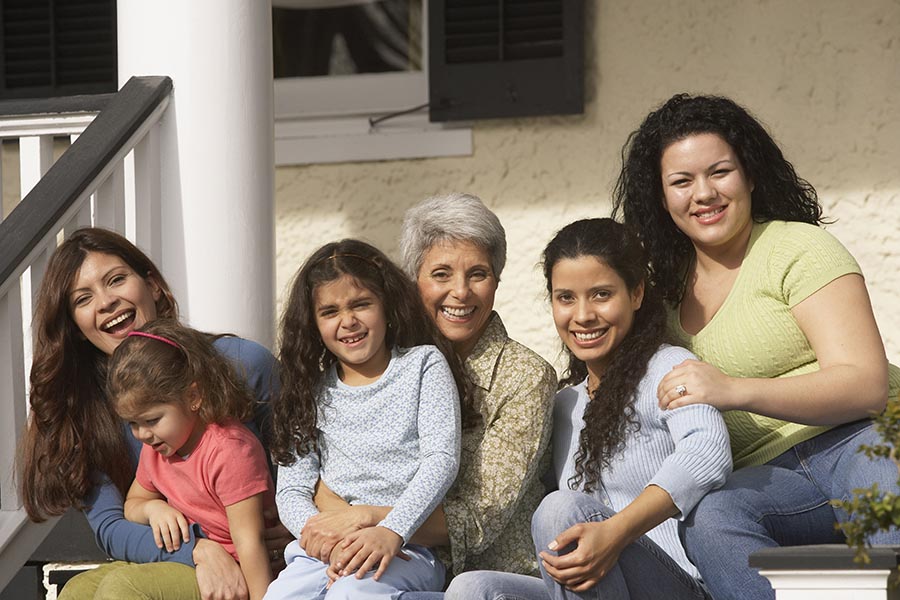 Multi-generational Latina family sitting outdoors on house porch steps, smiling, Adobe Stock.