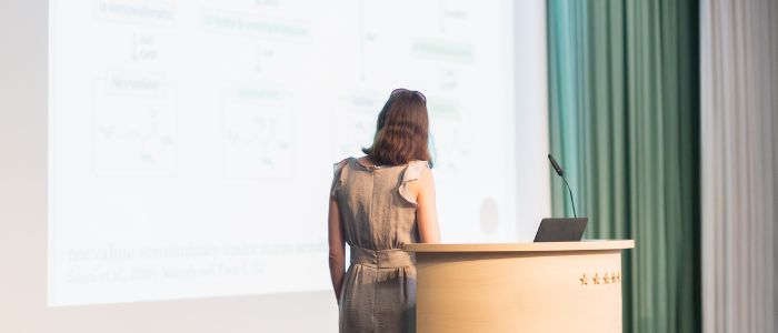 Woman at podium, back to the audience, facing a screen on the wall, giving a presentation; image credit Canva.